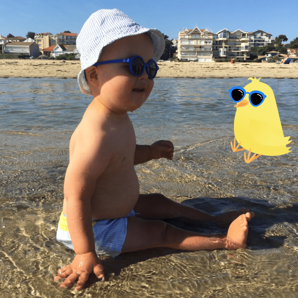 Un enfant portant un chapeau et des lunettes de soleil, assis au bord de l'eau sur une plage. Une illustration d'un poussin avec des lunettes de soleil est visible à côté de l'enfant