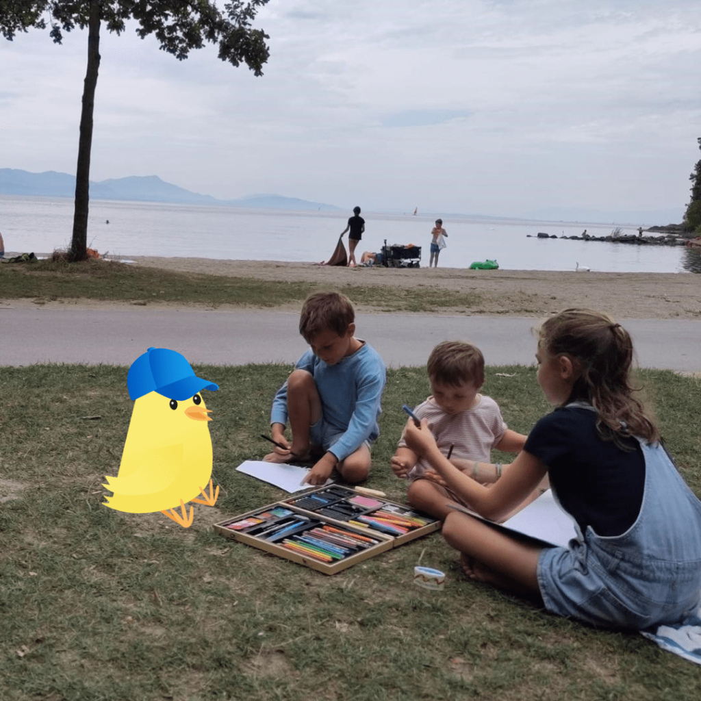 Trois enfants assis sur l'herbe, dessinant avec des crayons au bord d'un lac. Une illustration d'un poussin avec une casquette bleue est visible à côté d'eux.